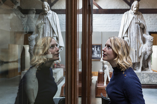 A woman looks in a cabinet at Watts Gallery. She and her background are reflected in the glass
